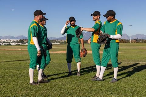 Baseball team huddling on sunny field wearing green uniforms and black caps discussing strategy