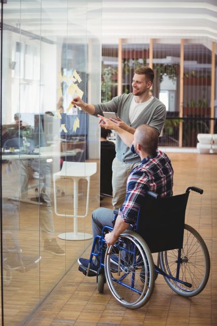 Handicap businessman assisting colleague while sticking adhesive notes on glass window in creative office