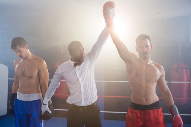 Referee raising hand of victorious boxer while the defeated opponent stands in the boxing ring. Ideal for use in sports articles, fitness blogs, motivational content, and advertisements related to boxing, sportsmanship, and athletic training.