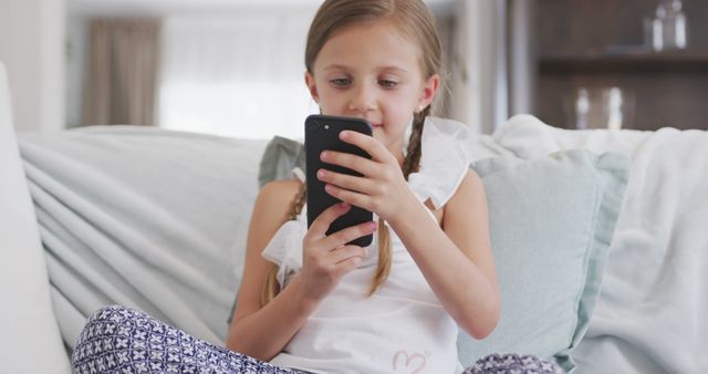 A young girl with blonde hair sitting on a white couch using a smartphone. She looks focused on the device, engaging with technology in a comfortable home environment. Ideal for concepts of childhood, digital lifestyle, at-home leisure, or modern communication.