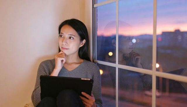 Asian woman sitting by window holding tablet, thinking at dusk with city reflections