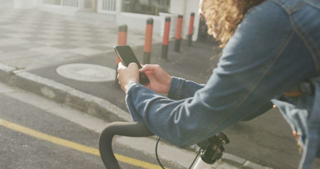 This image features a person riding a bicycle and using a smartphone in an urban street setting. The individual is wearing a denim jacket, holding a mobile phone in both hands, and the bicycle's handlebars are visible in the foreground. The scene suggests multitasking, daily life, and modern communication practices. Ideal for topics related to urban transportation, online connectivity, technology distractions, and cycling safety.