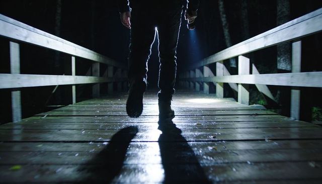 Backlit figure walking on wet forest footbridge at night holding flashlight beam