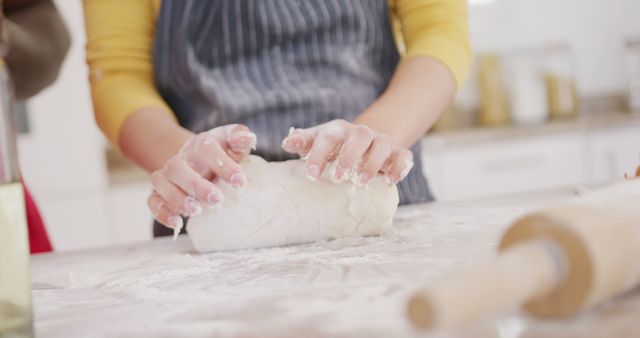 Person in yellow shirt kneading dough on floured kitchen countertop. Ideal for content related to home baking, cooking tutorials, recipe blogs, or kitchen-related products. The close-up focus on hands emphasizes the tactile process of preparing homemade bread or pastries.