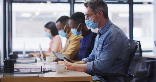 Color photo of several office workers sitting at a shared workspace, all wearing face masks. They are engaged with laptops and tablets, reflecting a professional environment adhering to pandemic safety guidelines. Ideal for articles on workplace safety, COVID-19 adaptations, or remote working tools.