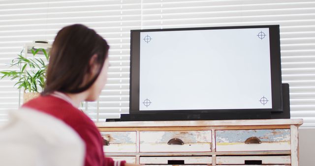 Person seated in living room watching TV displaying a calibration screen with crosshairs. Room features a worn wooden drawer and indoor plant. Ideal for topics related to technology setup, home electronics maintenance, and relaxation at home.