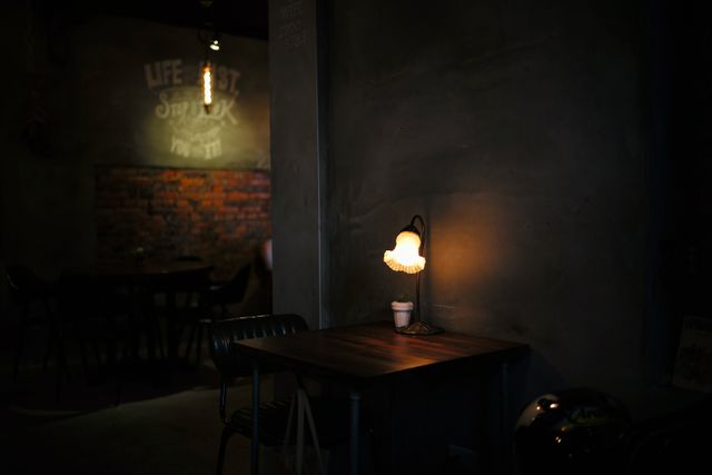 A cozy corner in a dimly lit café featuring a wooden table lit by a vintage table lamp, a single coffee cup, and warm light creating an inviting atmosphere. Ideal for use in designs related to intimacy, calm ambience, relaxation, cafes, and vintage decor settings. Perfect for blog posts about coffee culture, calm environments, or small business marketing materials.