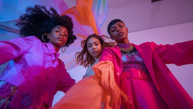 Low-angle trio of models reaching, posing in vibrant fuchsia blazers with flowing drapes