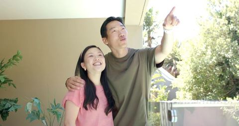 Asian couple enjoying sunny balcony on home patio, man pointing toward view, smiling