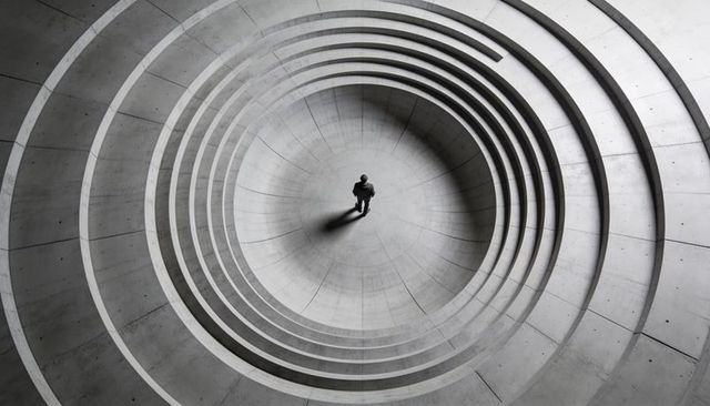Minimalist overhead view of lone man in concentric concrete rings casting long shadow