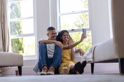Young couple sitting on floor taking selfie by sunlit windows in cozy modern home
