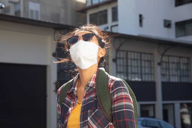 Biracial woman out and about in the city streets during the day, wearing sunglasses and a face mask for protection against covid 19 coronavirus, standing in a city street, looking up