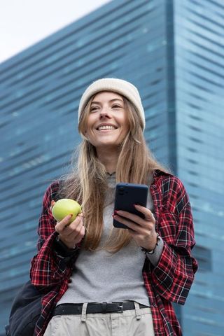 Young woman smiling with smartphone and green apple near modern glass office tower urban commute