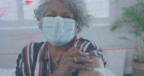 Senior woman wearing surgical mask showing vaccine bandage while sitting in clinic