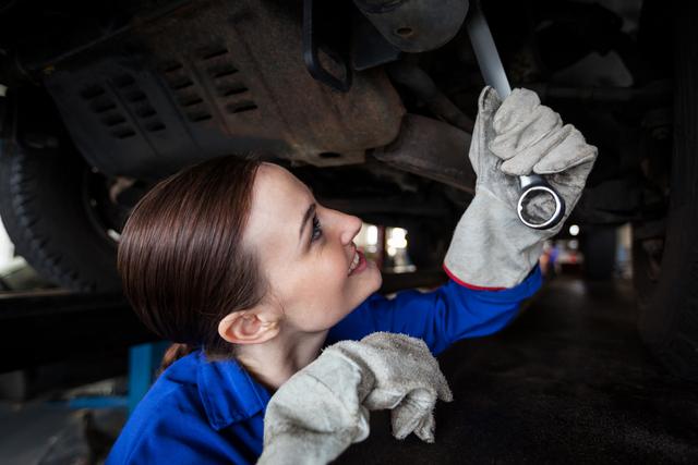 Female mechanic working on the undercarriage of a vehicle in a repair garage. She is wearing protective gloves and is focused on her task, showcasing her skills and professionalism. This image can be used for promoting auto repair services, highlighting female empowerment in technical fields, or educational purposes related to automotive training programs.