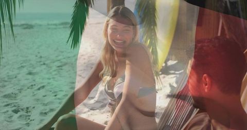 Laughing woman on tropical beach hammock with surfboard, sunlit couple vacation romance