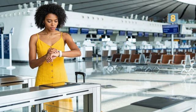 Young woman checking smartwatch at airport gate wearing yellow dress with carry-on luggage