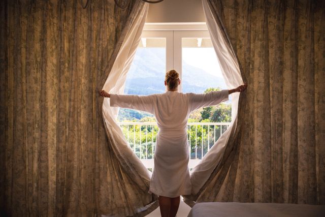 Caucasian woman wearing bathrobe, revealing the curtains, looking through the window. spending free time at home.