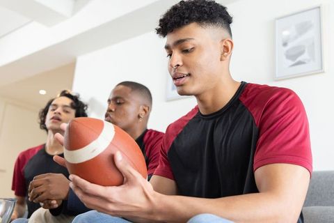 Multiracial young men examining football on couch preparing for game night and team bonding