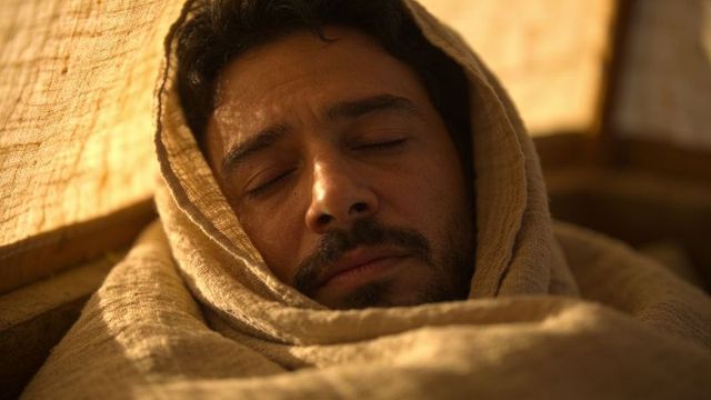 Calm middle eastern man wrapped in tan woven blanket sleeping inside narrow wooden shelter