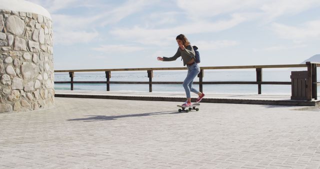 Young woman skateboarding on paved path by ocean, enjoying bright sunny day. Ideal for use in marketing materials related to healthy lifestyles, outdoor activities, beachside recreation, and youthful energy. Could also be used in travel and tourism advertisements or sports content.