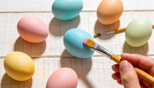Hand painting pastel easter eggs with brush on white wooden planks, spring craft scene