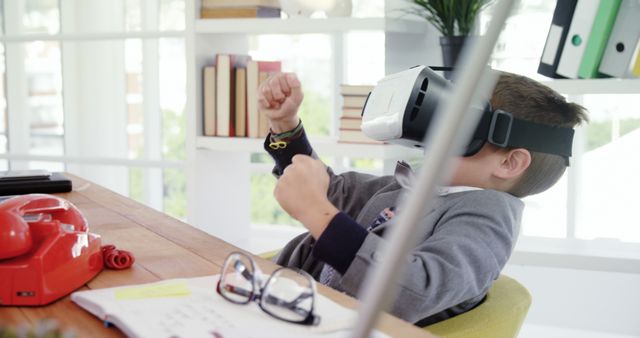 A boy in casual clothes wears a virtual reality headset and raises his fists in excitement, sitting at a wooden desk in a modern home office with shelves of books and plants in the background. Ideal for use in technology promotions, educational materials, or articles discussing the impact of virtual reality on learning and child development.