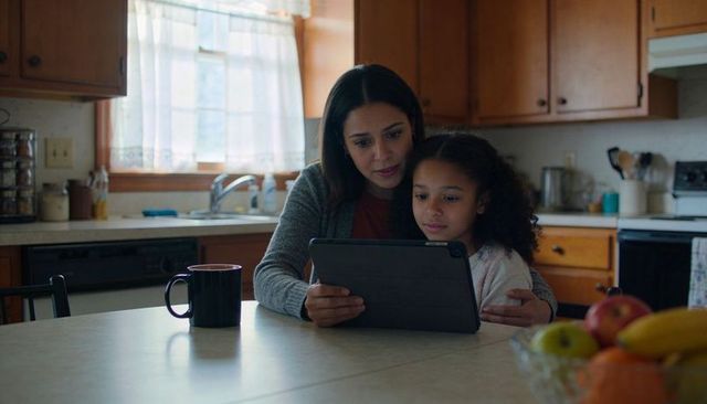 Mother guiding daughter using tablet at sunlit kitchen table for learning and bonding
