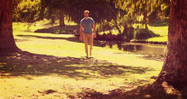 Man walking in a scenic park on a sunny day. Large trees casting shadows, and a serene stream runs in the background. Ideal for promotions related to outdoor activities, relaxation, wellness, or nature-focused content.