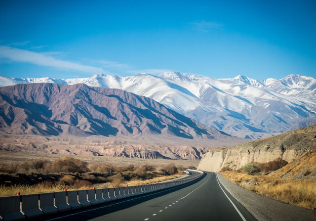 Scenic highway stretches through a breathtaking landscape featuring snow-capped mountains and arid desert surroundings. Image is perfect for travel blogs, adventure advertisements, and presentations focusing on road trips and natural beauty. Ideal for use in promotional materials and social media campaigns targeting outdoor enthusiasts and nature lovers.
