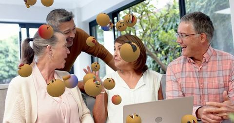 Mature friends laughing and collaborating around laptop with playful emoji overlays