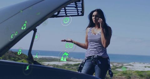 Indian woman talking on phone beside car with open hood on coast with eco holograms