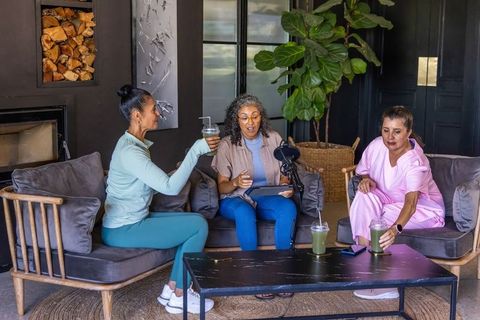 Diverse women recording casual podcast in cozy modern living room with smoothies together