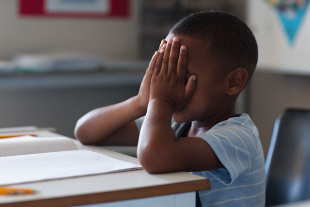 Sad african american elementary schoolboy covering face while sitting at desk in classroom. unaltered, education, studying, sadness, depression, failure, loneliness and school concept.