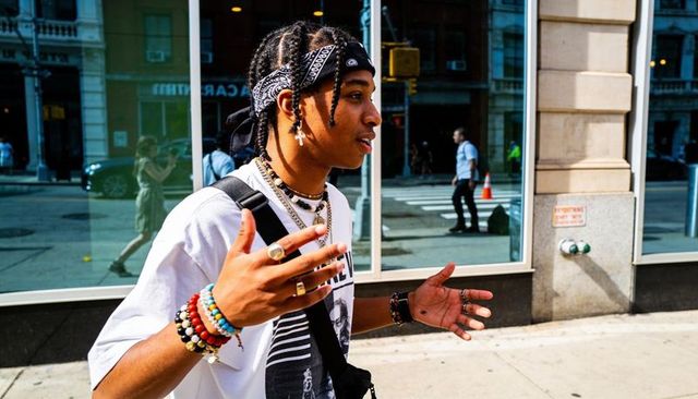 Man with braided hair and bandana walking and gesturing on urban sidewalk showing jewelry