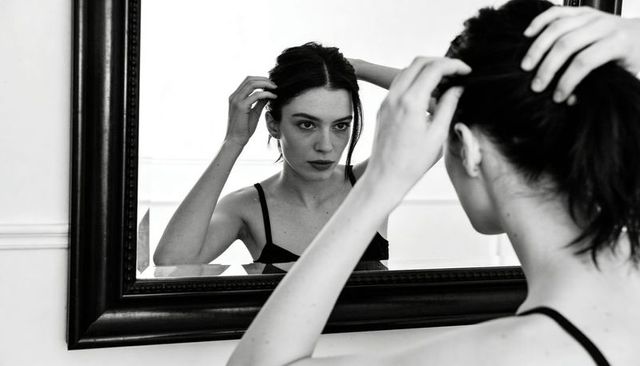 Contemplative woman adjusting hair in dark-framed mirror monochrome minimalist portrait