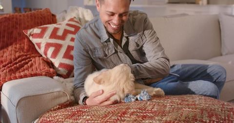 Smiling man cuddling golden retriever puppy on cozy sofa with rope toy