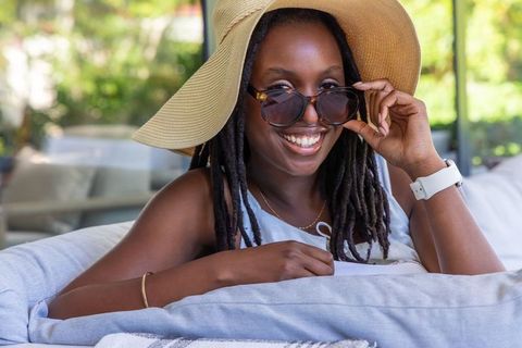 African american woman relaxing on patio sofa wearing straw hat and sunglasses, smiling