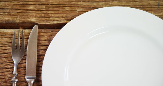 A fork and knife are placed beside an empty white plate on a wooden table, with copy space. The setting suggests preparation for a meal, emphasizing the importance of proper table setting.