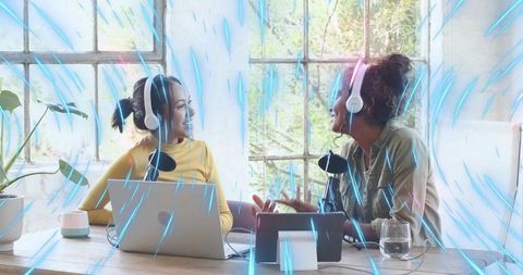 Recording podcast cohosts chatting and collaborating with laptops and headsets in sunlit studio