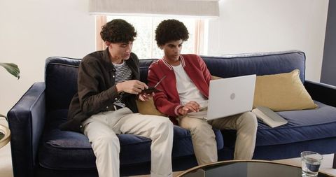 Two young men sitting on blue velvet sofa using laptop and smartphone in bright living room