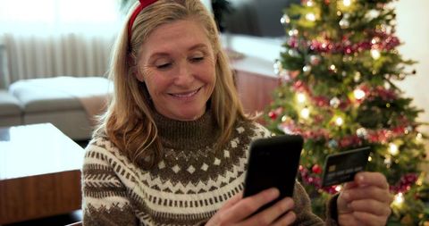 Joyful woman shopping online by christmas tree with smartphone