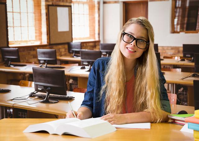 This image captures a smiling blonde teenage girl studying in a classroom setting. She is wearing glasses and writing on a paper, with a stack of books and a computer in the background. Perfect for use in educational blogs, school websites, study guides, and promotional materials for academic institutions.