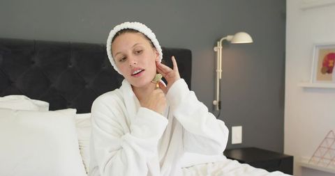African american woman using jade facial roller on neck in cozy white robe bedroom