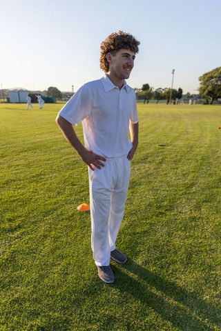 Young cricketer standing hands on hips on sunlit practice field in white kit
