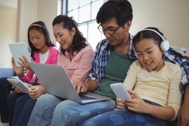 Family spending quality time together with various technological devices in living room. Father using laptop, mother with tablet, children with smartphones and headphones. Perfect for materials on modern family life, technology use, digital age, and home lifestyle.