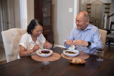 Senior asian couple sharing cozy home dinner with chopsticks and red wine at dining table