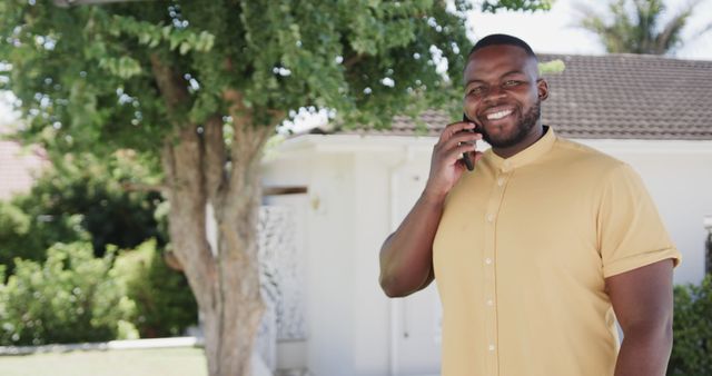 Man wearing casual clothing smiling and talking on smartphone outside house with tree in background. Ideal for commercial use related to communication, real estate, happy moments, and outdoor lifestyle.