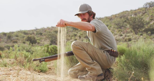 Kneeling caucasian male survivalist holding hunting rifle, examining sandy terrain of wilderness. exploration, travel and adventure, survivalist in nature.