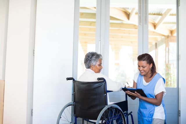 This image depicts a senior woman in a wheelchair consulting with a female caregiver in a home environment. The caregiver is holding a clipboard and smiling, indicating a supportive and caring interaction. This image is ideal for use in healthcare, elderly care, and home care service promotions. It can also be used in articles or brochures about patient care, disability support, and senior living.